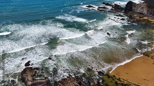 Wide aerial drone shot moving over large rolling waves and a rugged rocky beach with towering cliffs at Praia da Amália, Costa Vicentina, Portugal. Showcasing the powerful ocean meeting dramatic coast