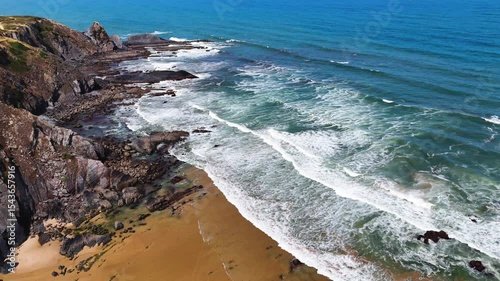 Wide aerial drone shot moving over large rolling waves and a rugged rocky beach with towering cliffs at Praia da Amália, Costa Vicentina, Portugal. Showcasing the powerful ocean meeting dramatic coast