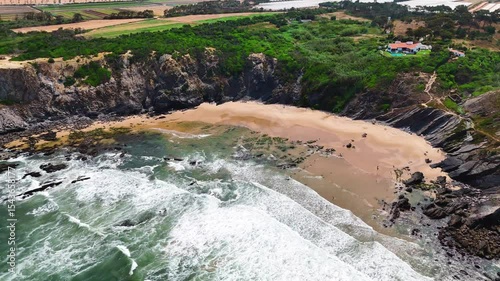 Wide aerial drone shot moving over large rolling waves and a rugged rocky beach with towering cliffs at Praia da Amália, Costa Vicentina, Portugal. Showcasing the powerful ocean meeting dramatic coast