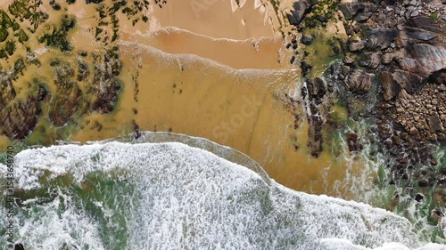 Top-down aerial view of ocean waves crashing onto the sand at Praia da Amália, Costa Vicentina, Portugal. Captured in slow motion, showing a dramatic coastal scene.