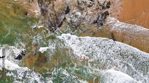 Top-down aerial view of ocean waves crashing onto the sand at Praia da Amália, Costa Vicentina, Portugal. Captured in slow motion, showing a dramatic coastal scene.