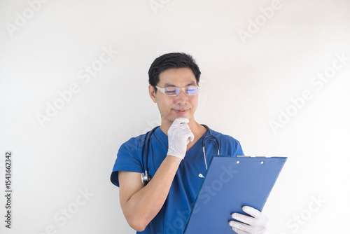 Asian male healthcare worker wearing blue scrubs holding a blue clipboard and looking at it thoughtfully