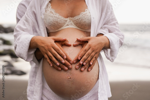 Happy couple holding hands on belly at the beach - Family, parenthood and maternity concept 