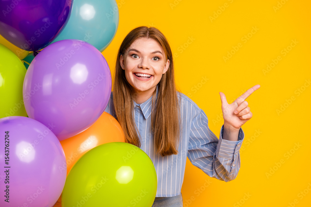 Fototapeta premium Cheerful young woman pointing and posing with colorful balloons against a vibrant yellow background in casual attire
