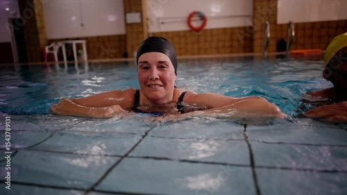 Female athlete wearing a black swim cap takes a break from her swimming practice, leaning on the pool's edge and smiling towards the camera