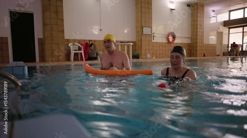 Senior couple engaging in water aerobics, supporting body movement with foam noodles during low impact workout within indoor swimming pool environment
