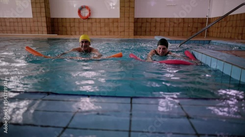 Senior couple performing gentle aqua aerobics in indoor swimming pool, engaging in low impact water exercise for rehabilitation, fitness, and wellness during retirement years