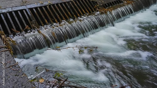 A flowing stream of water cascades over a wooden barrier. The water is clear with some debris floating on the surface. The scene is tranquil and natural.