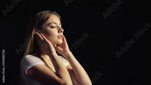 Young Caucasian woman with long brown hair, holding her hands to her ears, in a dark setting. The scene conveys a sense of contemplation or introspection.