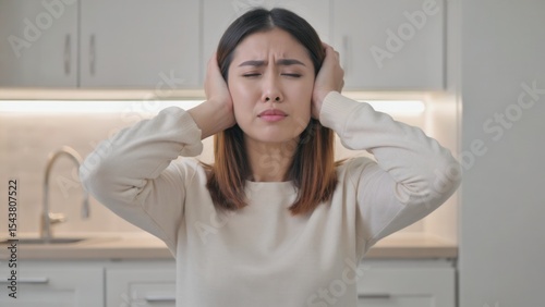 A young Asian woman with shoulder-length brown hair covers her ears with her hands. She appears distressed in a modern kitchen setting.