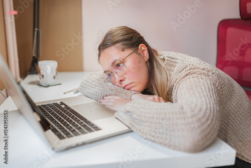 Tired woman resting chin on arms beside laptop, blank expression.	Emotionally flat mood, arms folded on desk, head resting near laptop with eyes open, suggesting exhaustion or burnout in quiet work