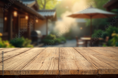 Empty rustic wooden table with copy space in a sunny summer garden. In the background barbecue grill garden umbrella and cozy holiday cabin. Outdoor party, grilling time and summer relaxation concept.