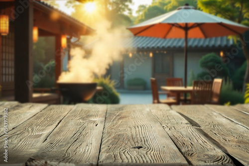 Empty rustic wooden table with copy space in a sunny summer garden. In the background barbecue grill garden umbrella and cozy holiday cabin. Outdoor party, grilling time and summer relaxation concept.