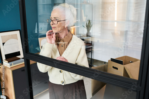 Senior Caucasian woman standing by glass wall in modern office setting with moving boxes visible in background suggesting retirement transition
