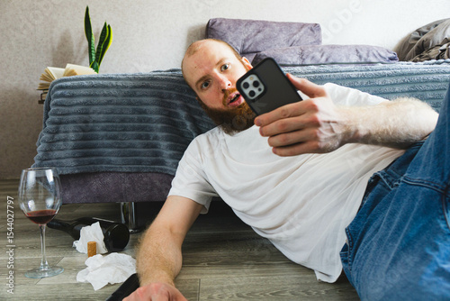 Surprised bearded man lying on the floor, holding smartphone and staring at screen with shocked expression. Messy scene with wine glass, bottle, and tissues suggests emotional or dramatic situation.

