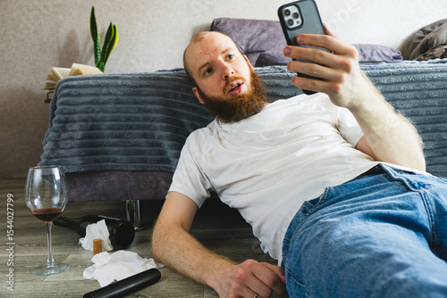 Surprised bearded man lying on the floor, holding smartphone and staring at screen with shocked expression. Messy scene with wine glass, bottle, and tissues suggests emotional or dramatic situation.

