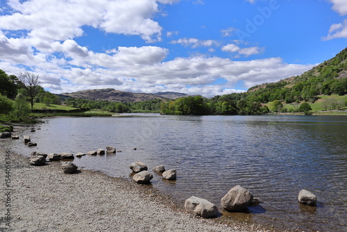 Shoreline of Rydal Water with scattered rocks, calm water and surrounding green hills under a partly cloudy sky. Lake District, England, UK. photo