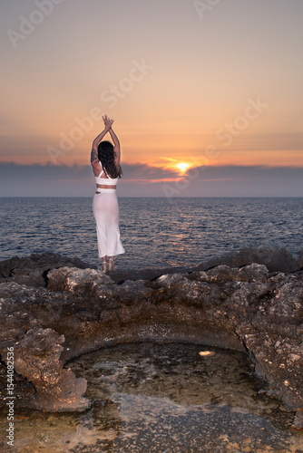 Fit girl in a semi-transparent white skirt watches the sunset on a cape in Menorca