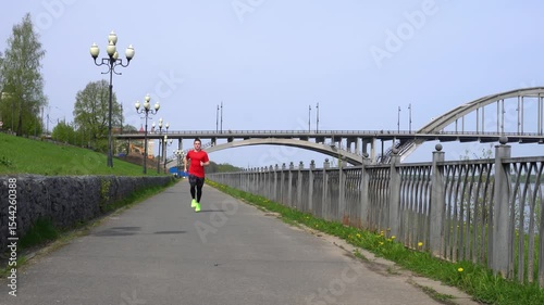 A young man in a red sports T-shirt runs in an urban environment near the bridge. A teenager performs cardio training