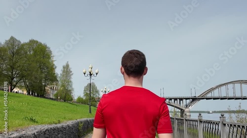A guy in a red T-shirt walks along the embankment against the backdrop of a bridge in Rybinsk, preparing for a running workout