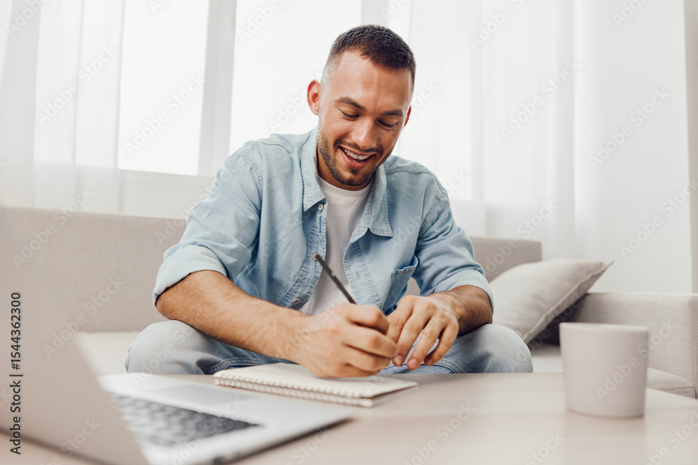 Fototapeta premium Smiling man engaged in creative work, writing in a notebook, wearing casual attire, surrounded by a bright, modern workspace with soft natural light.