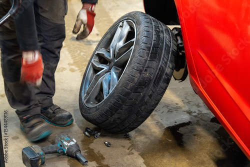 Winter to Summer Tire Change Service with Removed Wheel Leaning on Red Vehicle in Garage Workshop. Seasonal Tire Replacement Process Showing Winter Tread Pattern on Dismounted Tire with Hydraulic Jack