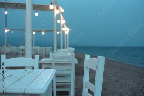Under the ethereal glow of sea lanterns, whitewashed tables anticipate coastal gatherings, evoking Aegean evenings and serene solitude