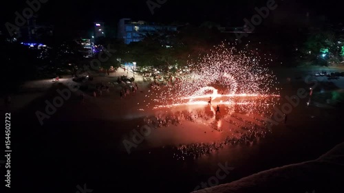 Nighttime beach spectacle featuring fire dancers performing with mesmerizing flames, creating luminous reflections on wet sand while tourists watch in awe during festive tropical celebration