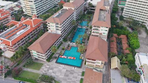 Elevated view of tourists swimming and playing in a large pool with inflatable water park features at a tropical resort surrounded by palm trees and hotel buildings