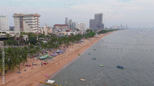 Relaxing tourists enjoying sandy Pattaya beach, framed by palm trees and urban high rise buildings along picturesque Thai coastline under bright sunny skies