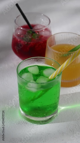 Three glass tumblers with red, yellow, and green lemonade on white background. Cold summer drinks with ice and condensation drops on glass surface. Refreshing for hot weather and restaurant