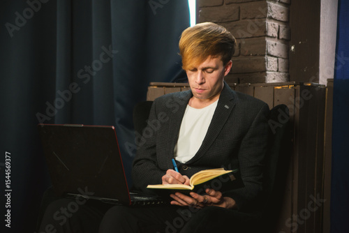 A focused young man sits with a laptop, writing in a notebook. He wears a blazer and t-shirt in a dimly lit room, concentrating on his work or studies.