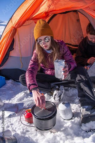 Woman cooking freeze-dried meal on portable stove at snowy mountain campsite. Winter outdoor cooking during alpine expedition
