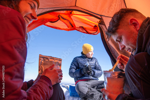 Winter camping in tent. Smiling hikers eat freeze-dried meals with snow-covered mountains in background