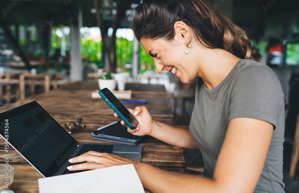 Fototapeta premium Smiling young woman using smartphone while typing on laptop, enjoying remote freelance work in cozy café environment, surrounded by modern tech tools and natural daylight