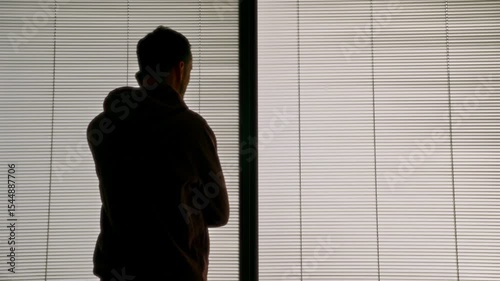 Young man with hoodie walking talking and listening to phone call in office. Businessman or student standing by apartment window speaking to smartphone. Silhouette with venetian blinds in background 