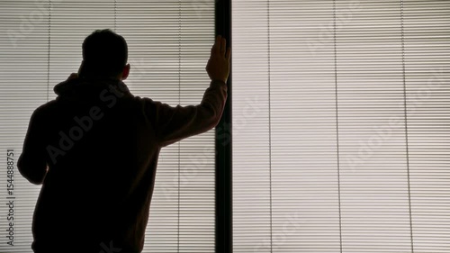 Man leaning on window with shutters or venetian blinds talking on the phone. Young businessman or student holding mobile phone speaking in office or apartment home. Business deal or important call