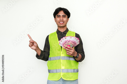 Happy Asian construction worker smiling while holding money and pointing sideways, isolated on white background. Perfect for salary, success, and promotion advertisement themes