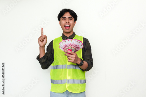 Happy Asian construction worker smiling while holding cash and pointing upward, isolated on white background. Great for themes about achievement, income, bonus, or financial success