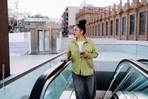 thoughtful latina woman on an escalator checks her banking app on a smartphone. She reviews transactions, manages finances, and prepares for online purchases while commuting in an urban setting