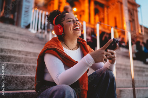 joyful hispanic woman wearing wireless headphones laughs while engaging with her smartphone. The city lights illuminate the urban scene, enhancing the digital experience of music and social media