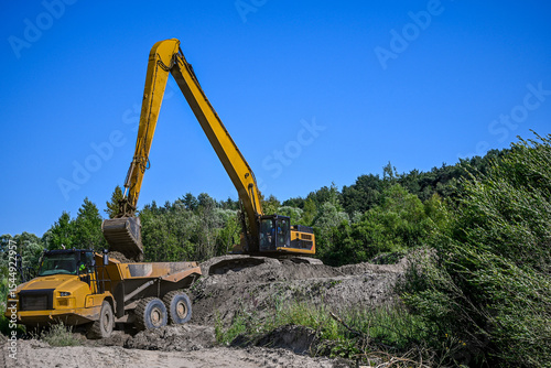 Excavator loading a dump truck with soil on a construction site near a forest, under a blue sky