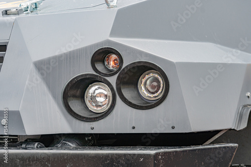 Close-up view of a gray, armored demining vehicles front bumper Three circular metallic elements in triangular formation, possibly lights or sensors, are fastened to the surface A flat section wit