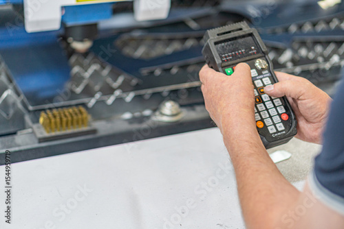 Person operating a handheld industrial controller, likely for machinery or equipment, in an indoor workspace with heavy machinery and artificial lighting