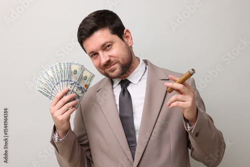 Greedy banker with dollar banknotes smoking cigar on grey background