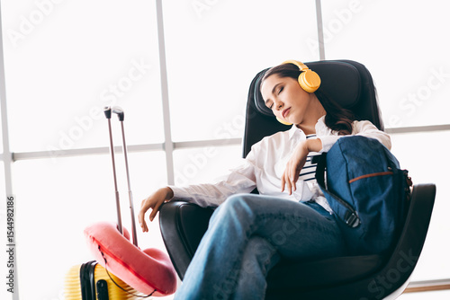 Tablou pe pânză Young female traveler resting at airport lounge with headphones and luggage, emb