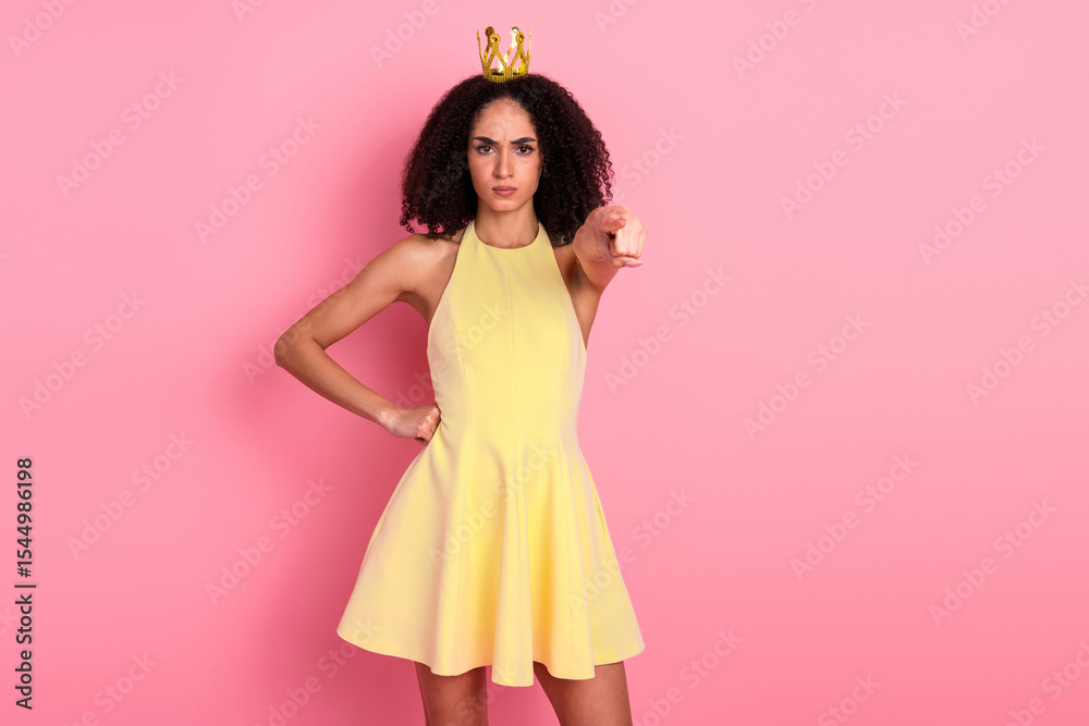 Young stylish woman with natural curls wearing a yellow dress and a crown pointing forward, set against a pink backdrop Young stylish woman with natural curls wearing a yellow dress and a crown pointing forward, set against a pink backdrop