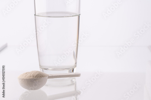 Collagen powder in a measuring spoon and a glass of water on a white background with reflection. Medicinal powder prepared for ingestion or dissolution in water.