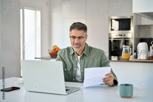 Middle aged mature man senior customer wearing glasses looking at laptop computer holding paper bill checking financial invoice or tax document making online banking payments working at home table.