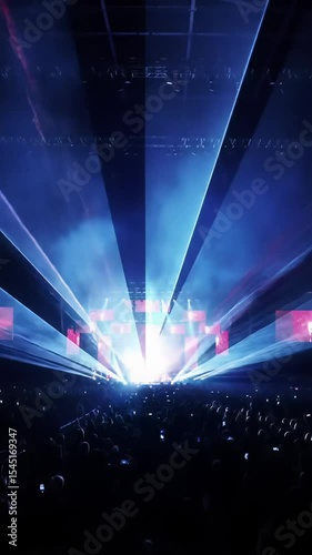 Slow crane video of a vast silhouetted audience in colossal arena. Intense blue/crimson spotlights, lasers, haze create a mesmerizing spectacle of vibrant light and deep shadow, exhilarating concert